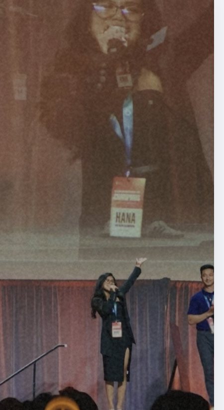 Ms. Hana speaking on stage to a large audience